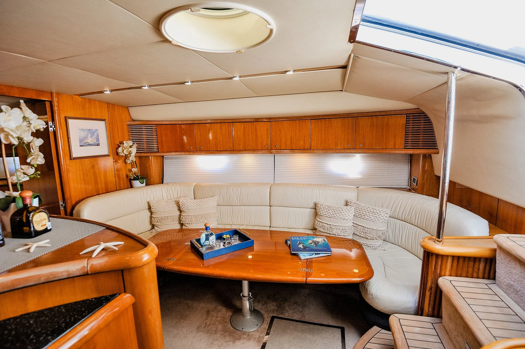 A cozy, wood-paneled lounge area on a boat or yacht with a curved cream-colored leather seating arrangement, a wooden table with magazines and bottled drinks, and decorative flowers on the side. The ceiling has a round light fixture and small recesse
