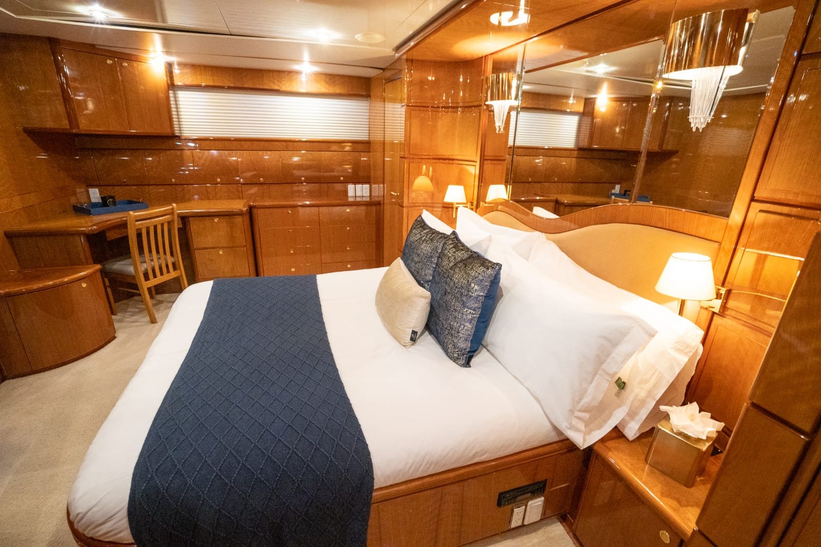 A bedroom with wood-paneled walls and a large bed with white linens, decorative pillows, and a blue quilted runner. The room has bedside lamps on on each side of the bed, a mirrored headboard, a small desk and chair, and a window with a closed blind.