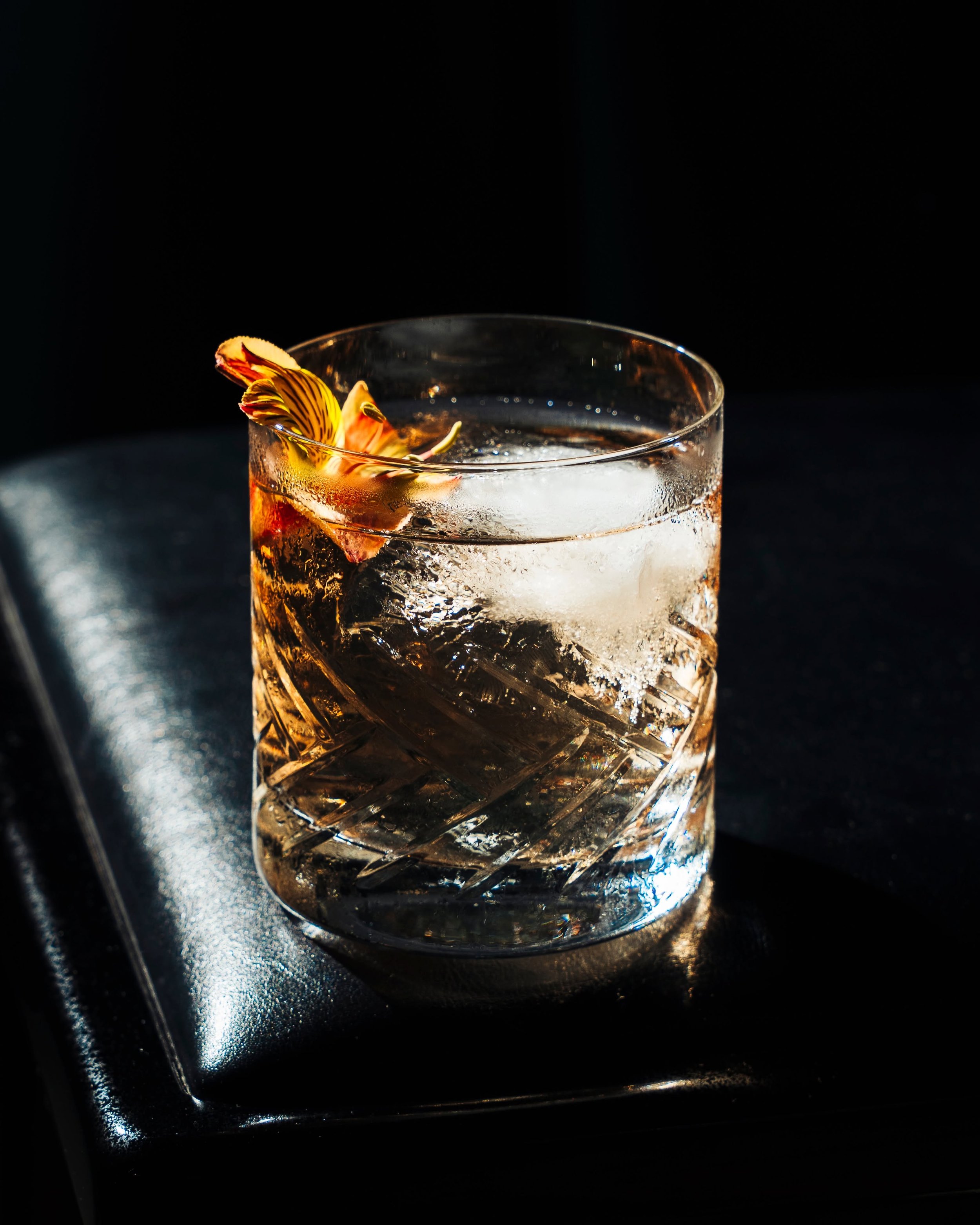 A close-up of a glass filled with a clear beverage and ice, garnished with a flower and a peel of citrus fruit, set against a dark background.