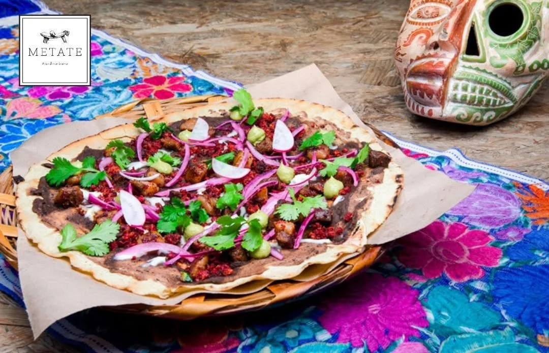 Mexican dish called Tostada with sliced red onions, cilantro, guacamole, and crumbled meat on a crispy tortilla, served on a colorful embroidered cloth with a decorative skull mask in the background.