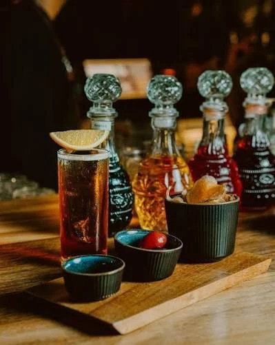 An assortment of flavored syrups and a tall glass of soda with a lemon wedge, along with small bowls of cherries, on a wooden serving board in a dimly lit setting.