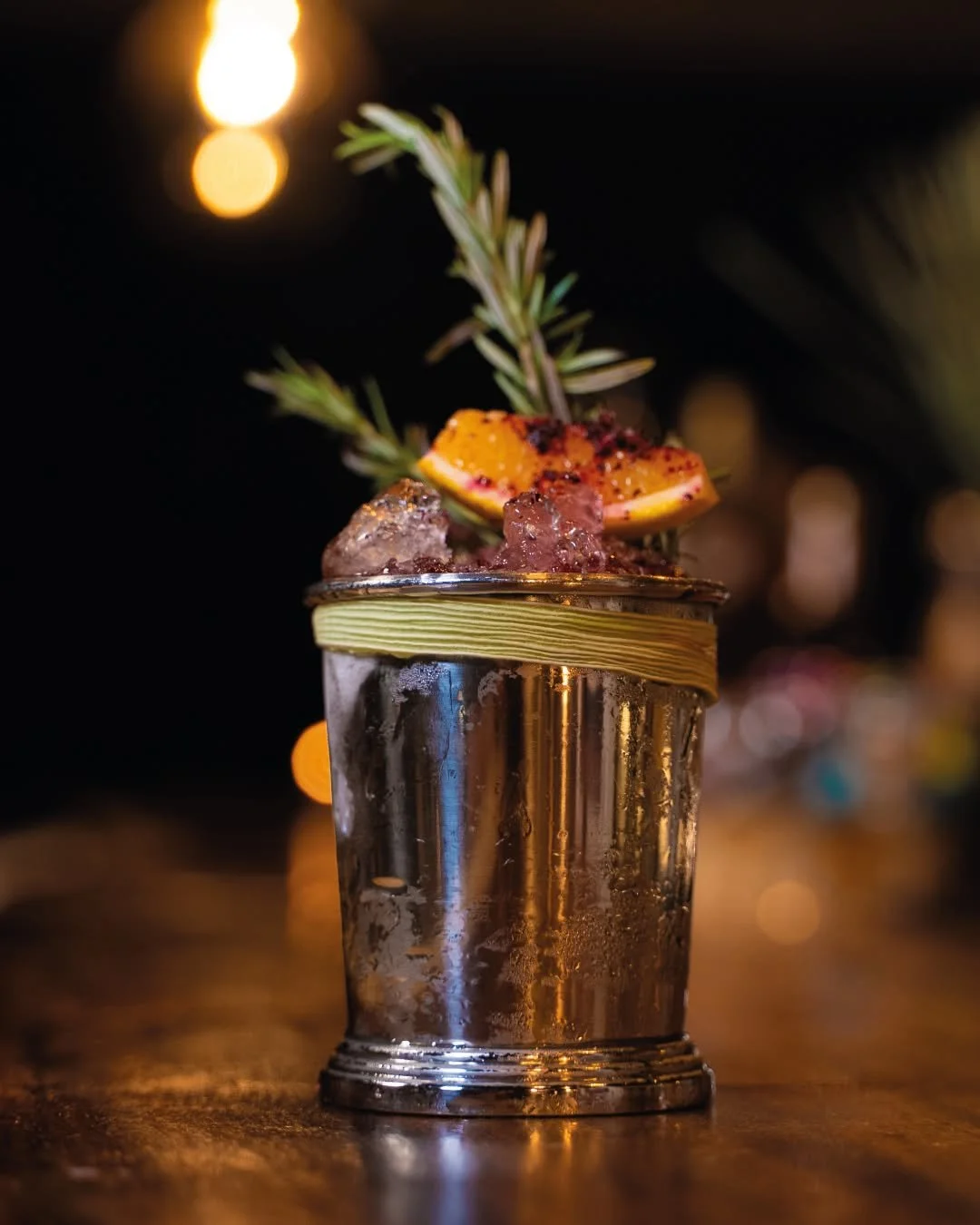 A cocktail served in a shiny metallic cup garnished with a sprig of rosemary and a lemon slice with black pepper, with ice cubes inside, on a wooden surface under warm, dim lighting.
