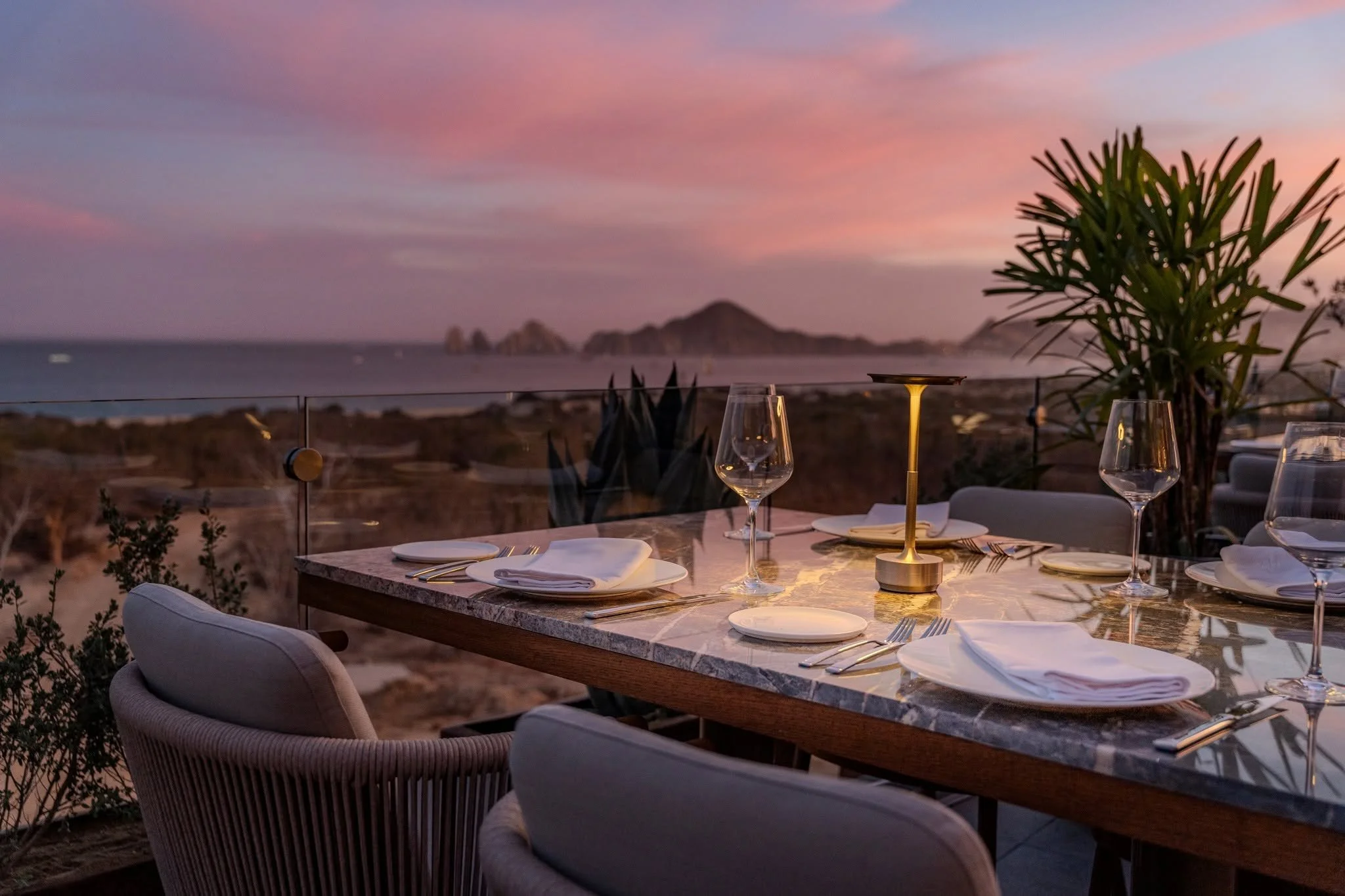 A restaurant table set for dining outdoors at sunset, with wine glasses, plates, and cutlery, overlooking a scenic view of the ocean and islands in the distance, with pink and purple clouds in the sky.