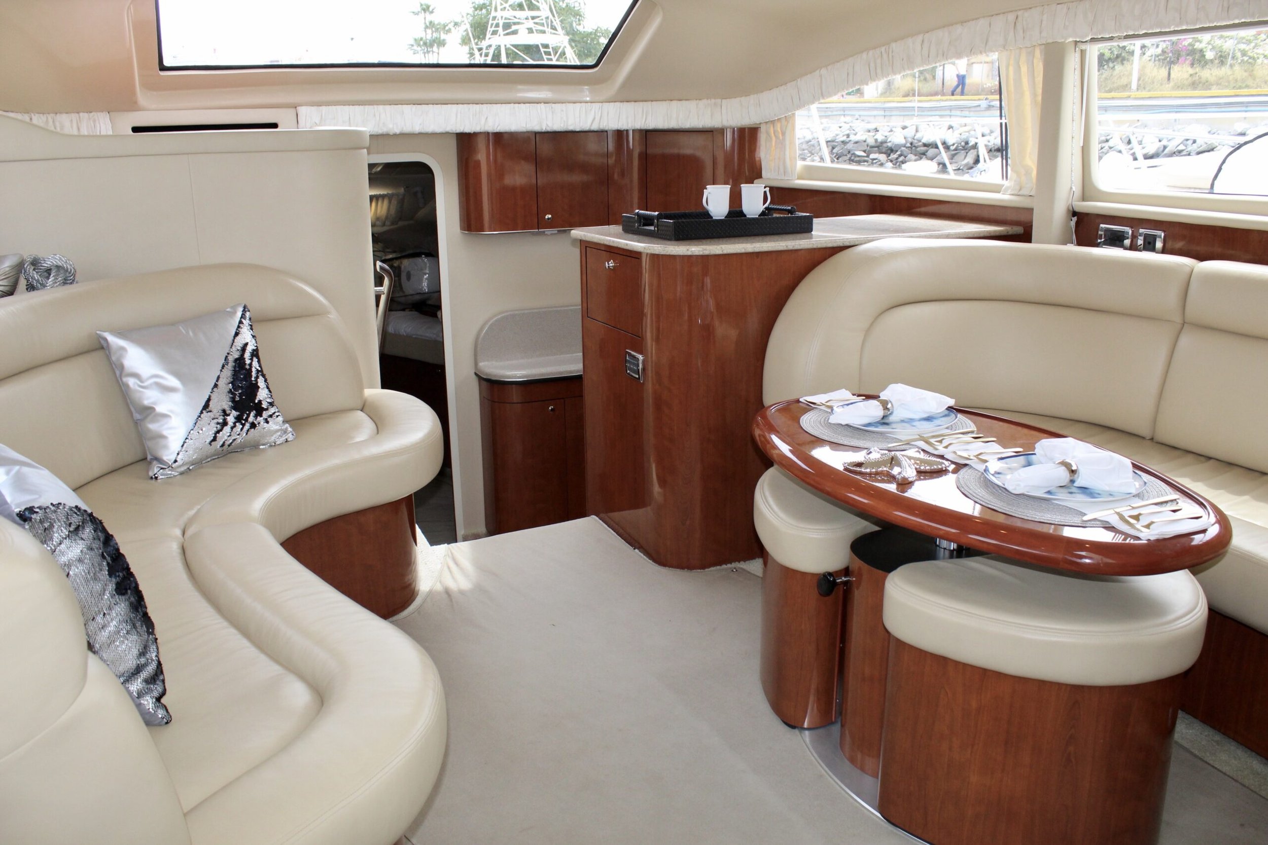 Inside a yacht or boat with beige leather seating, a dining table set with plates, napkins, and silverware, and a window showing an outdoor scene with railway tracks and a person walking.