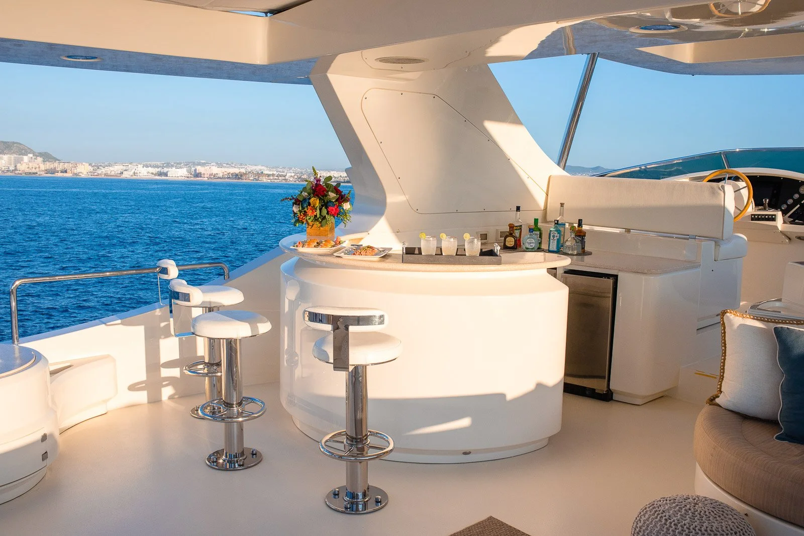 View of the deck of a yacht with a bar area, bar stools, a small refrigerator, and a floral centerpiece, overlooking the ocean with a distant shoreline under a clear sky.