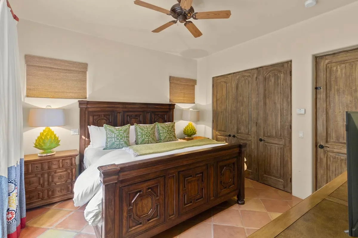 A bedroom with a wooden bed and matching nightstands, decorated with green pillows and matching lamps shaped like yellow artichokes. The room has a ceiling fan, terracotta tile flooring, and wooden closet doors.