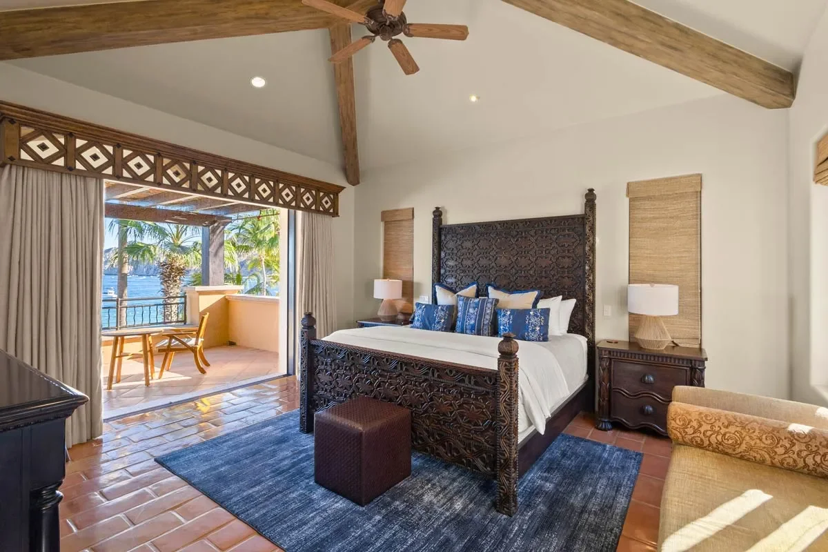 A bedroom with a large carved wooden bed, blue and white pillows, two nightstands with lamps, a dark blue rug, a small brown ottoman, and a sliding glass door opening to a balcony with a table and chairs overlooking palm trees and water.