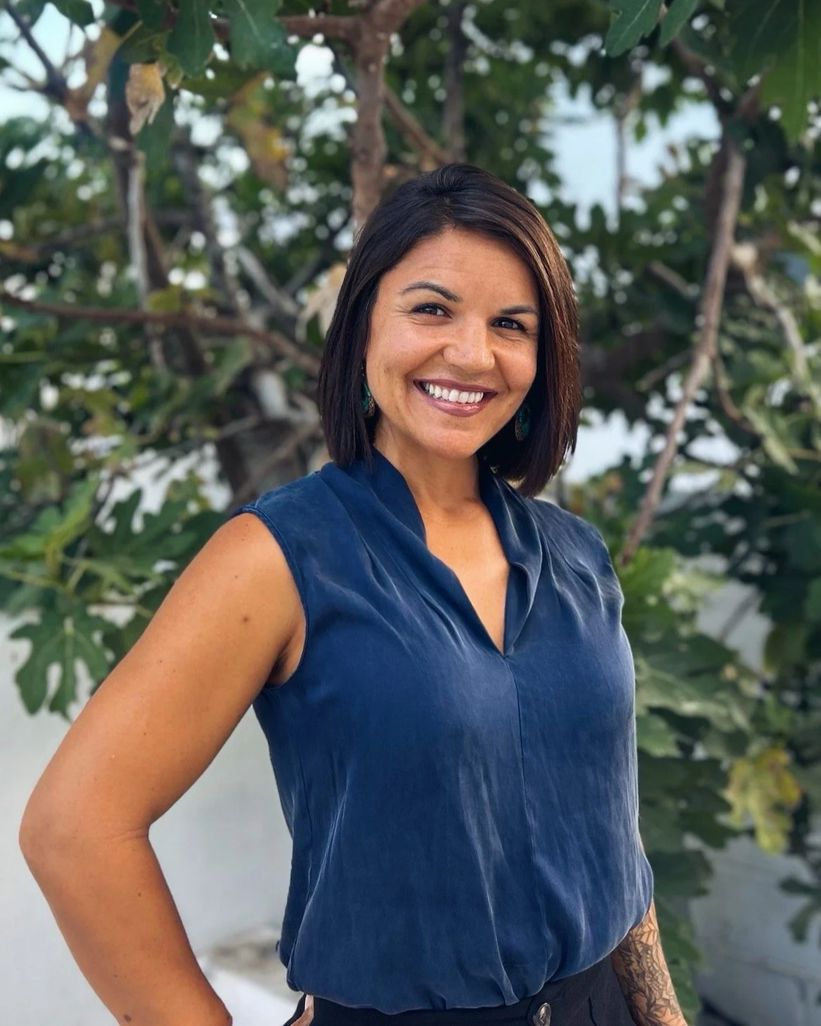 Smiling woman with shoulder-length dark hair wearing a sleeveless navy blue top standing outdoors in front of a leafy tree.