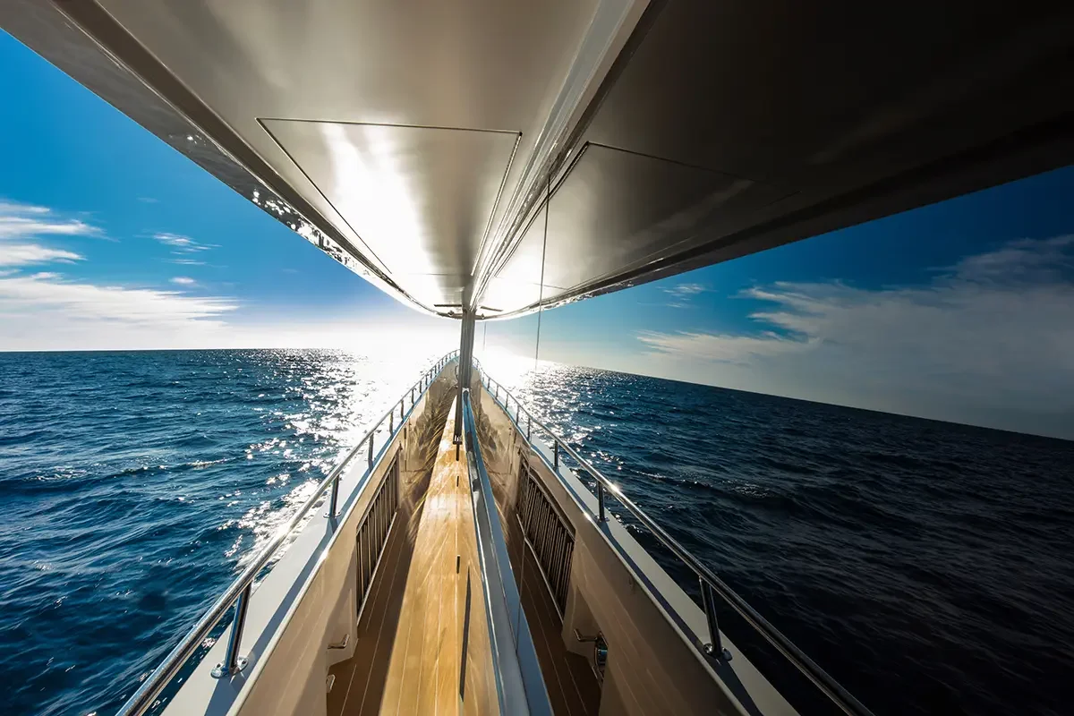 Underwater view from a boat showing the deck with railings, the ocean extending to the horizon, and the sky with scattered clouds, with the sun shining in the distance.