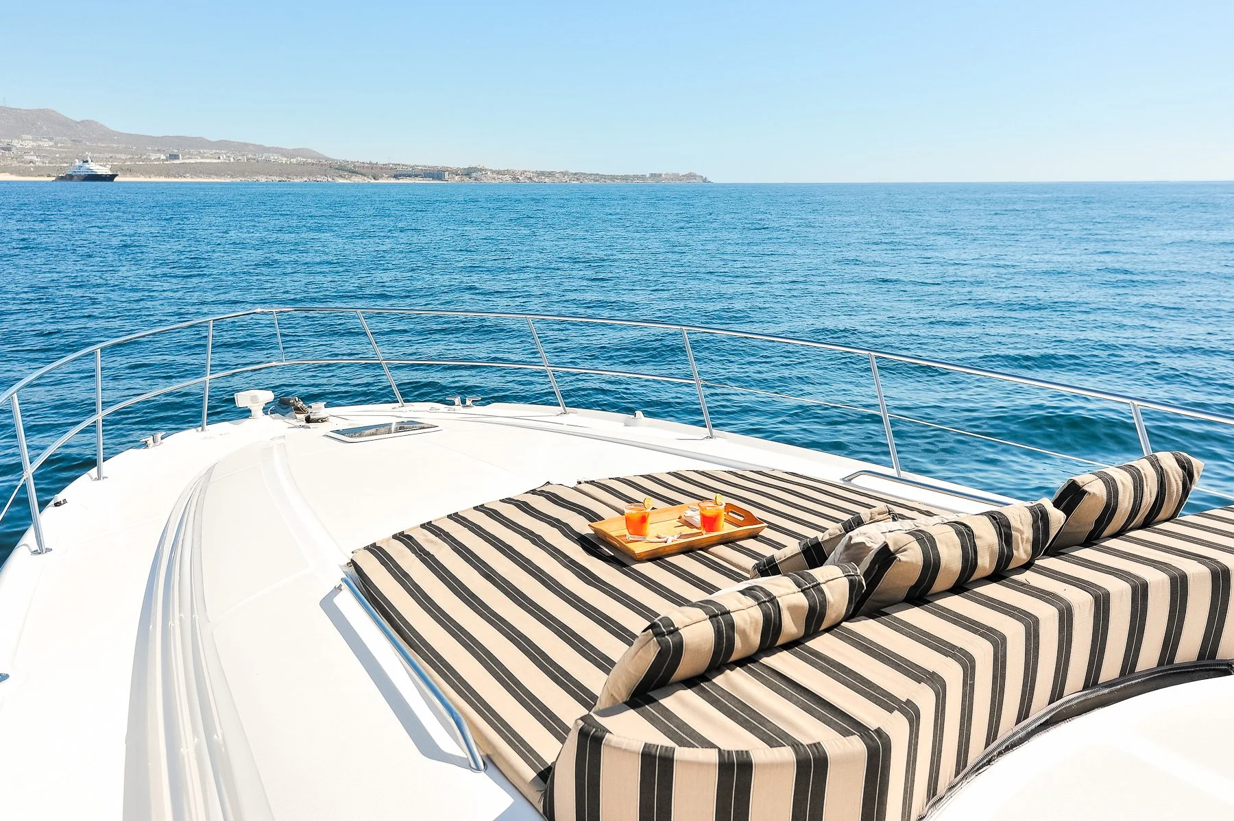 View from a yacht deck showing a striped cushioned lounge area with a tray of two drinks and snacks, overlooking calm blue ocean with a distant shoreline and another yacht in the background.