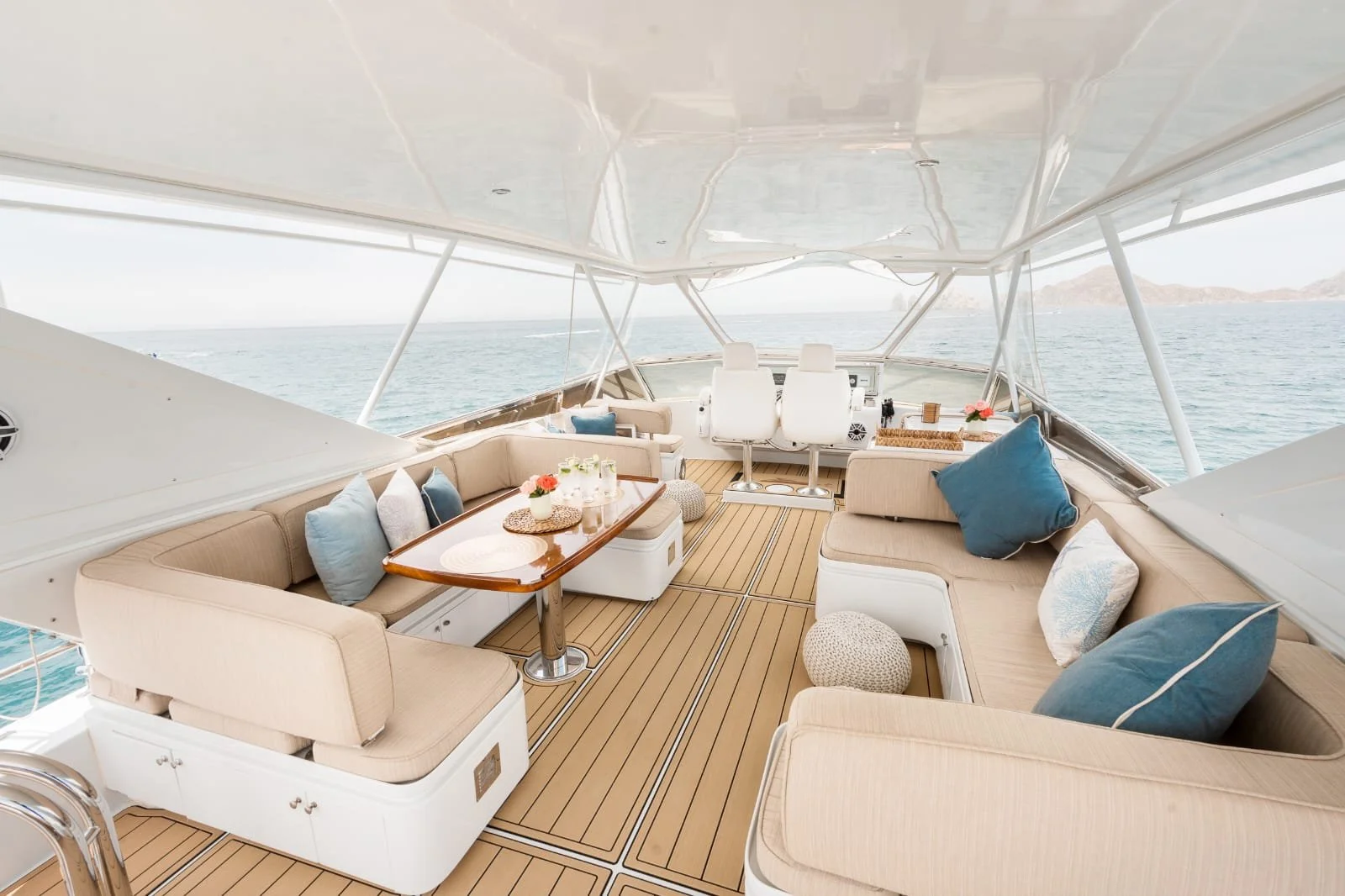 View of a luxury yacht's outdoor seating area with beige cushions and colorful pillows, wooden table, and ocean in the background.