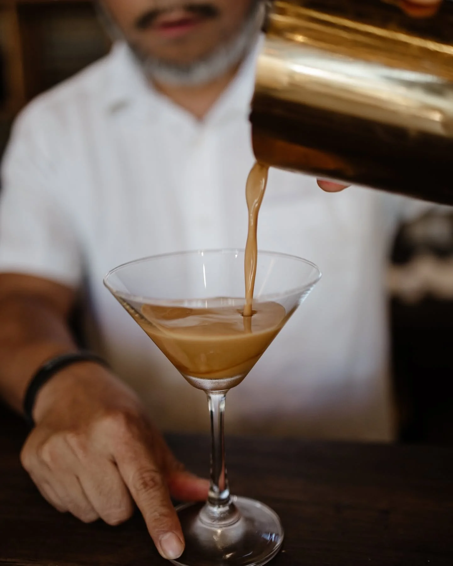 A man in a white shirt pours a brown cocktail from a shaker into a martini glass, with his hand and business ring visible.
