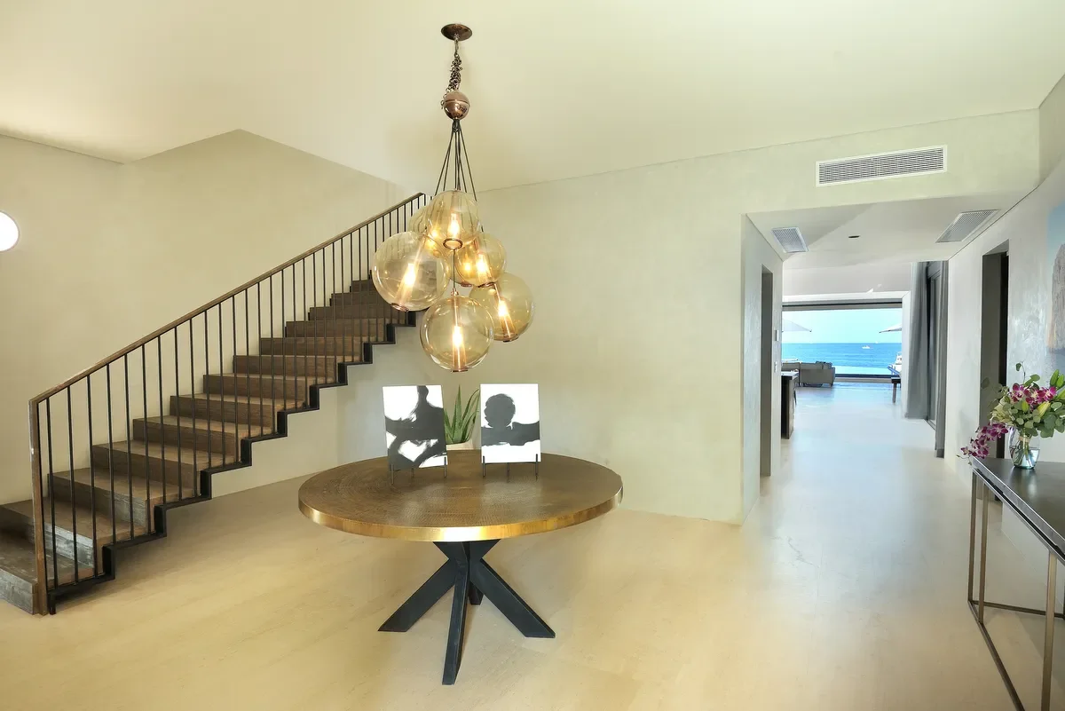 Modern living space with a wooden dining table, hanging glass pendant lights, staircase with black railing, and a view of the ocean through a hallway.