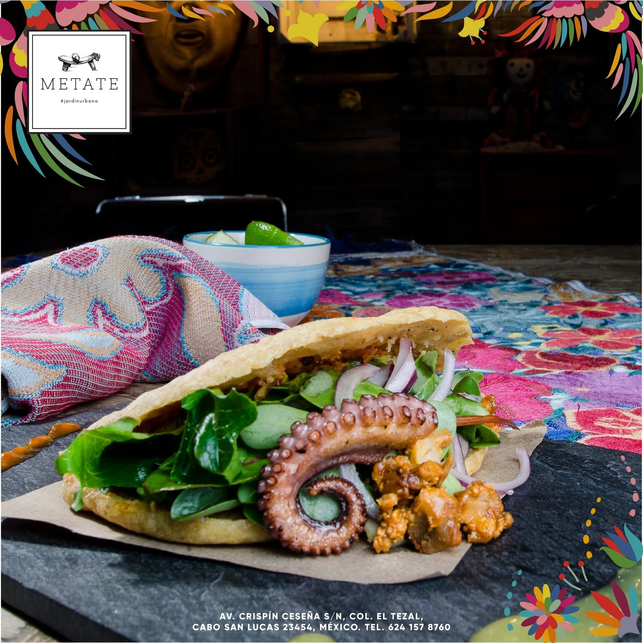 Close-up of a taco filled with lettuce, onions, and meat on a colorful, patterned cloth, with a bowl of lime slices and a breadstick in the foreground. The background shows a dark indoor setting with wooden shelves and decorations, and a framed logo that reads 'METATE' with a hashtag #jardinurbano.