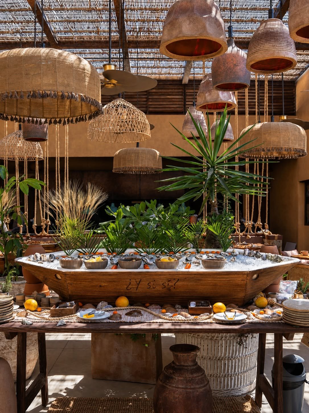 Decorative indoor setting with hanging wicker and clay lamps, tropical plants, and a wooden boat filled with bowls, fruits, and utensils, with the words "¿Y tú sí?" written on the boat.