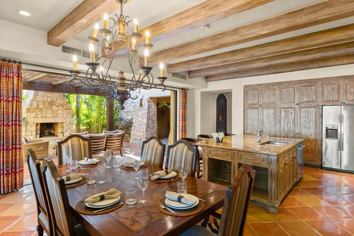 Interior of a dining room and kitchen area with a large wooden table set for a meal, a chandelier hanging above, wooden beams on the ceiling, a stone outdoor fireplace visible through sliding glass doors, and a stainless steel refrigerator in the kit
