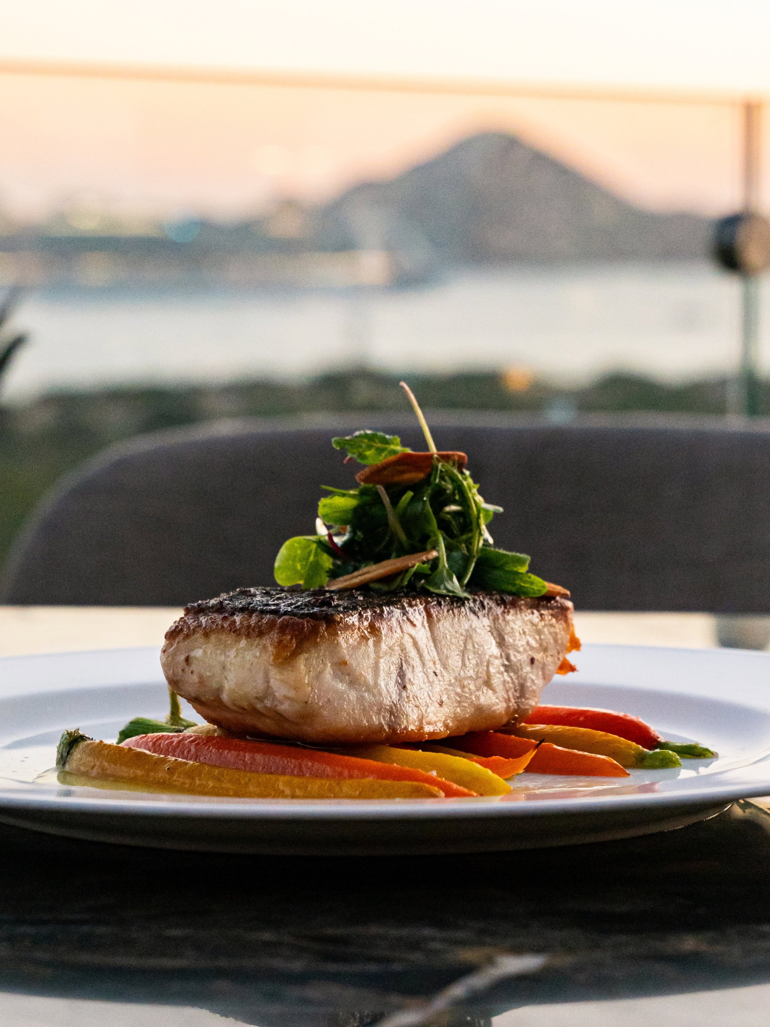 A plated dish featuring a cooked fish fillet topped with herbs and greens, served over a bed of assorted colorful roasted vegetables, with blurred mountain scenery in the background.