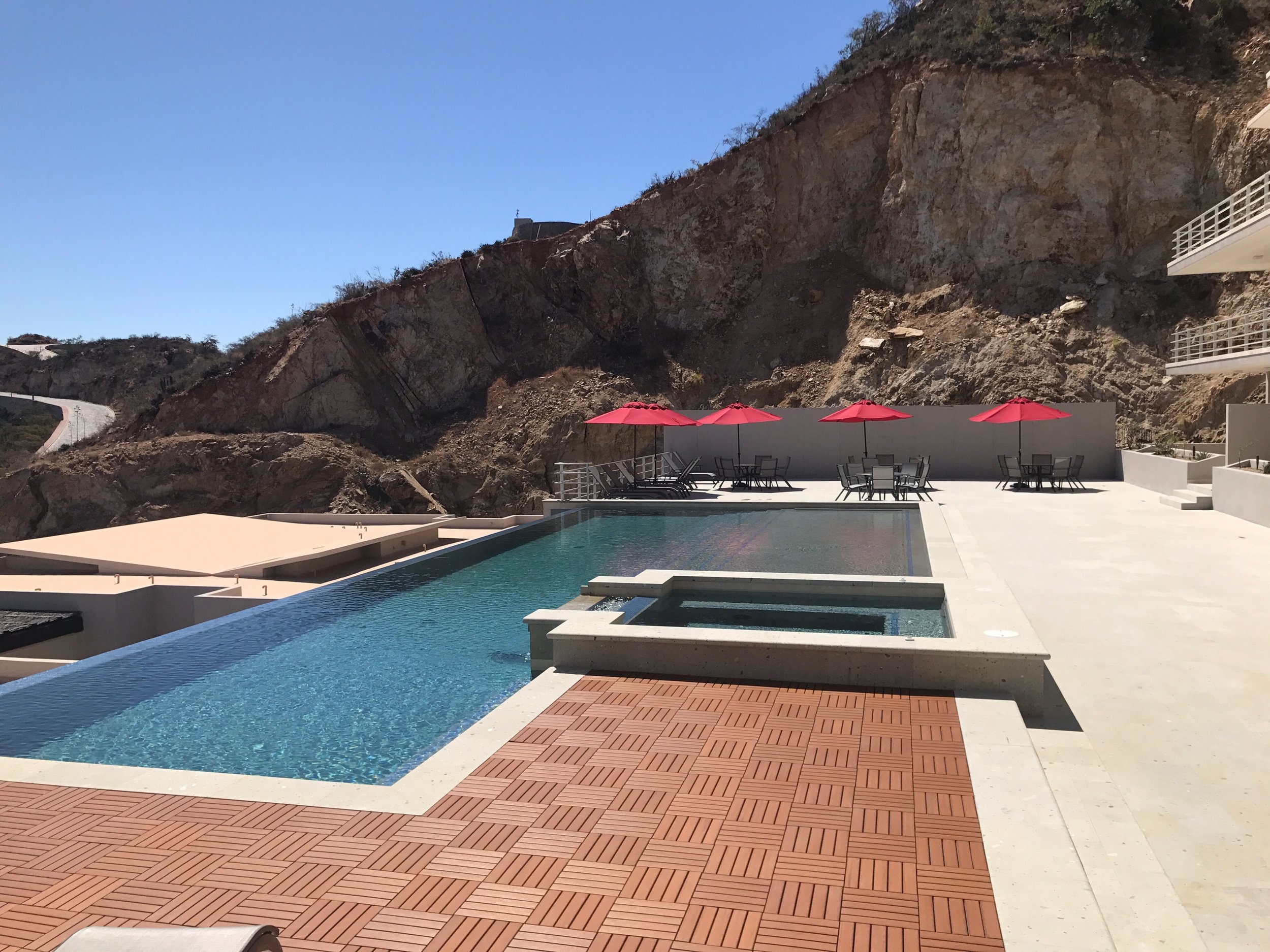 View of an outdoor swimming pool with red umbrellas and patio chairs, set against rocky hillside terrain and clear blue sky.