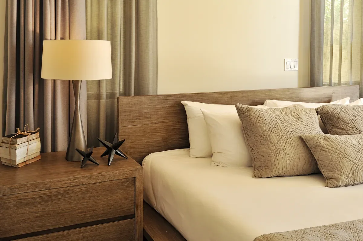 A bedroom with a wooden bed, beige and brown pillows, a wooden nightstand, a cream-colored lamp, and beige curtains.