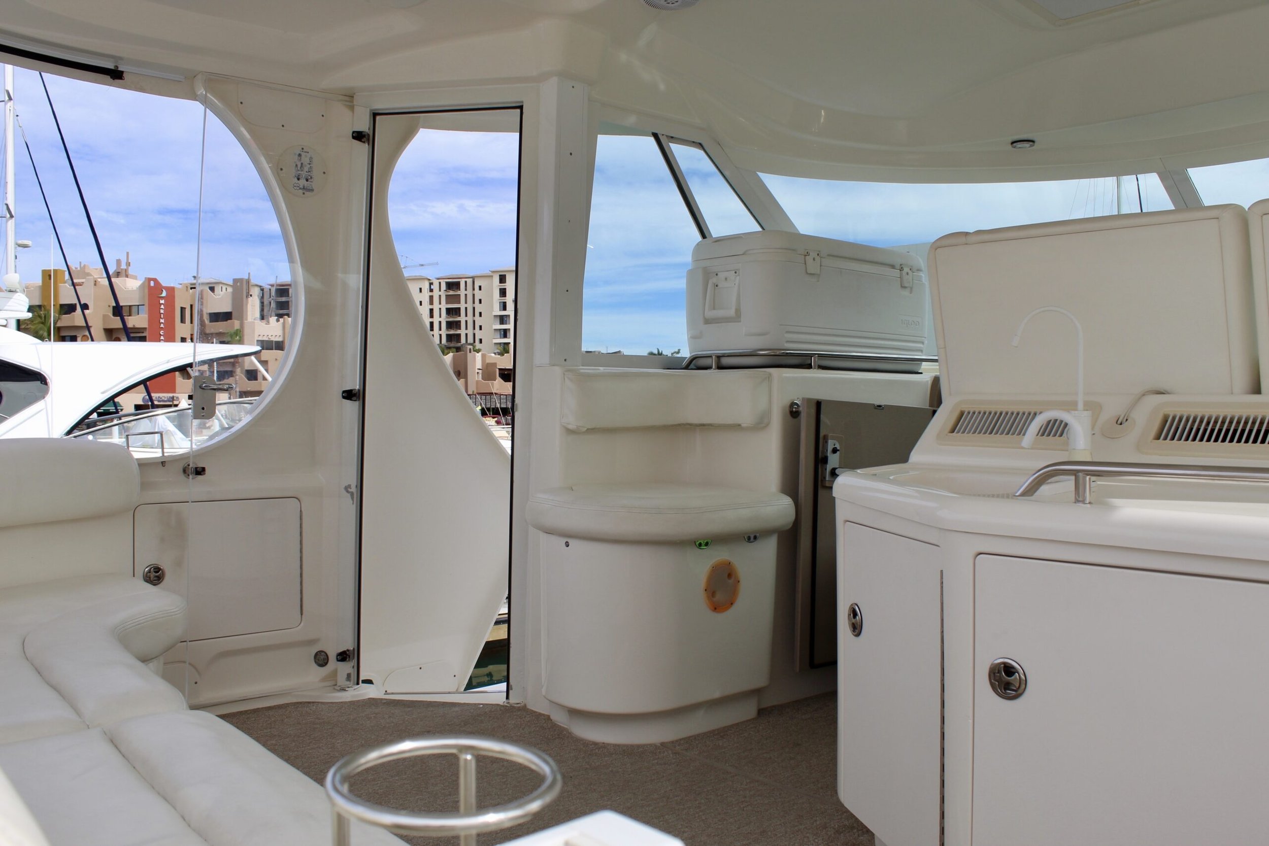 Interior view of a white yacht with seating, cabinets, and windows showing buildings and a cloudy sky outside.
