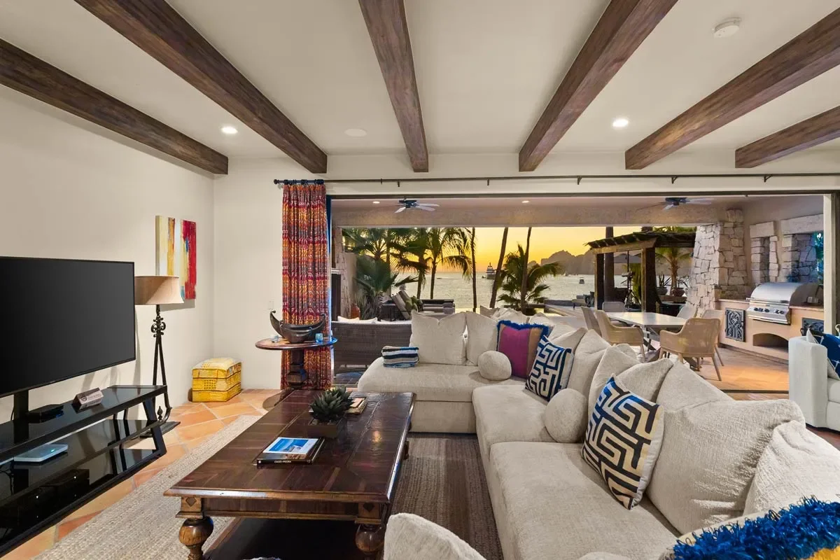 Living room with large beige sectional sofa, decorative pillows, a wooden coffee table, TV on a black stand, colorful artwork, a lamp, and a sunset view of a patio with outdoor furniture, palm trees, a grill, and the ocean in the background.