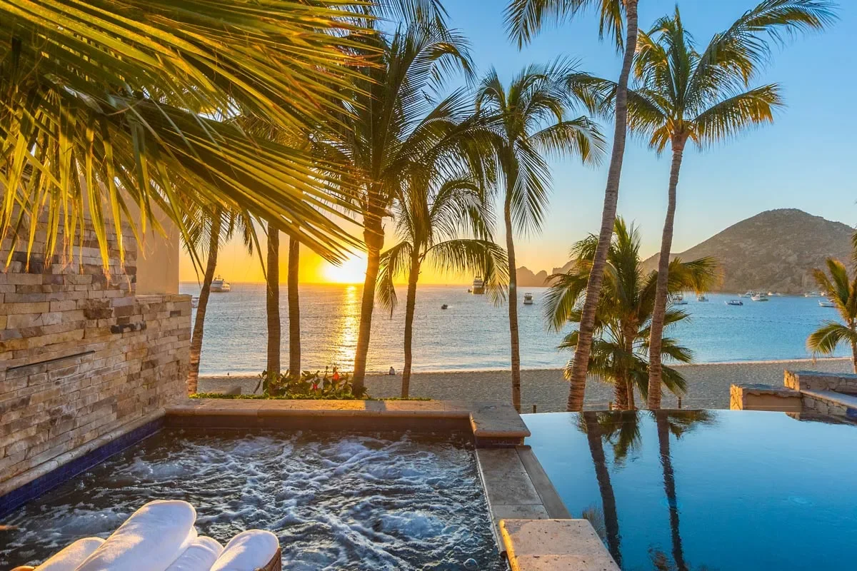 Sunset view from a poolside with palm trees and a beach in the background, boats on the water, and mountains in the distance.