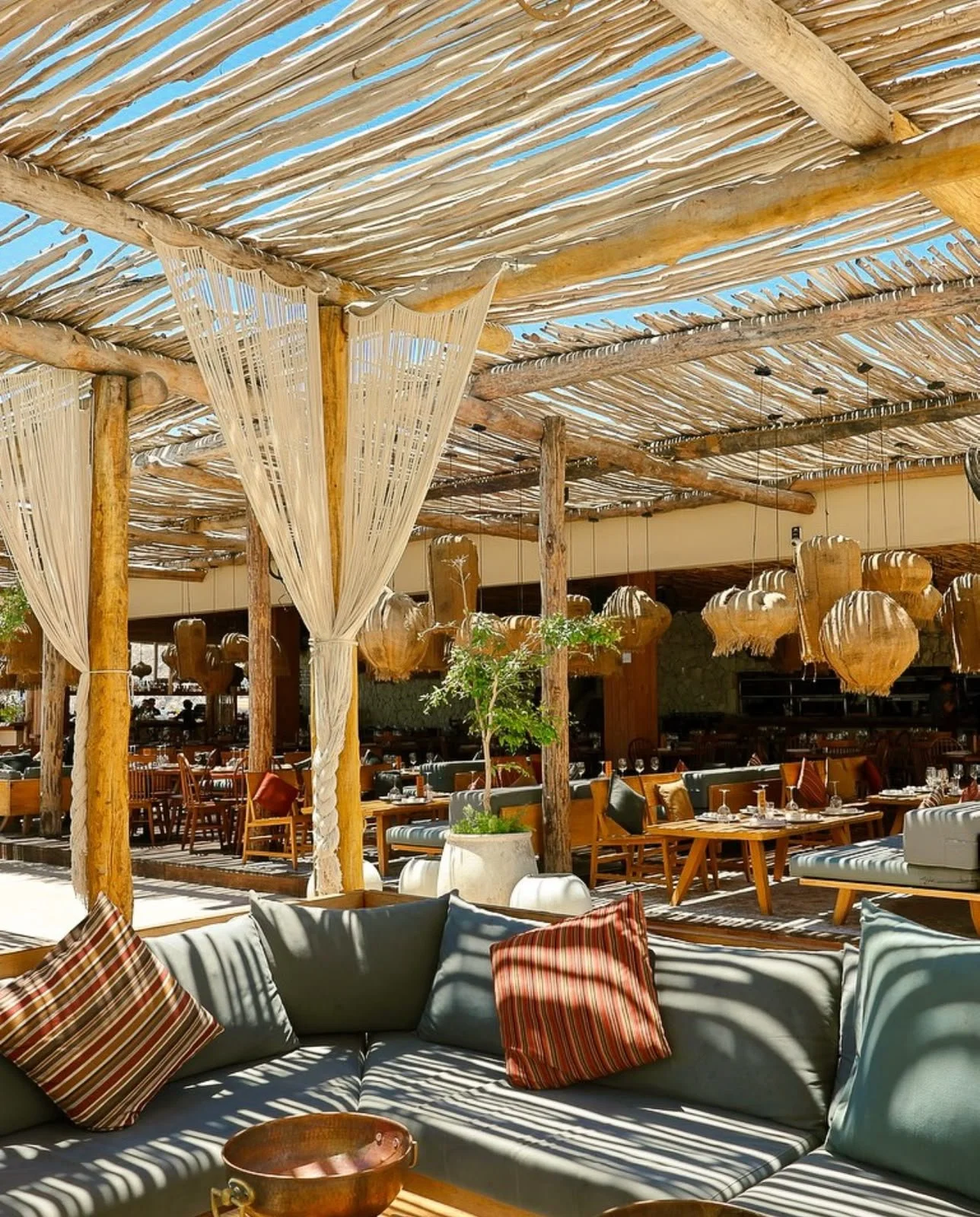Outdoor restaurant with wooden beams, fabric curtains, and hanging lamps, featuring tables, chairs, and a sofa with striped pillows under a thatched roof.