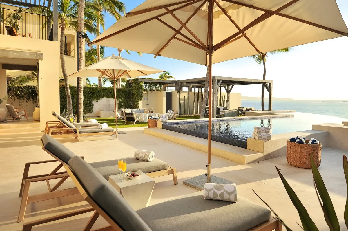 Luxury poolside area with lounge chairs, umbrellas, and an infinity pool overlooking the ocean, surrounded by palm trees and modern outdoor furniture.