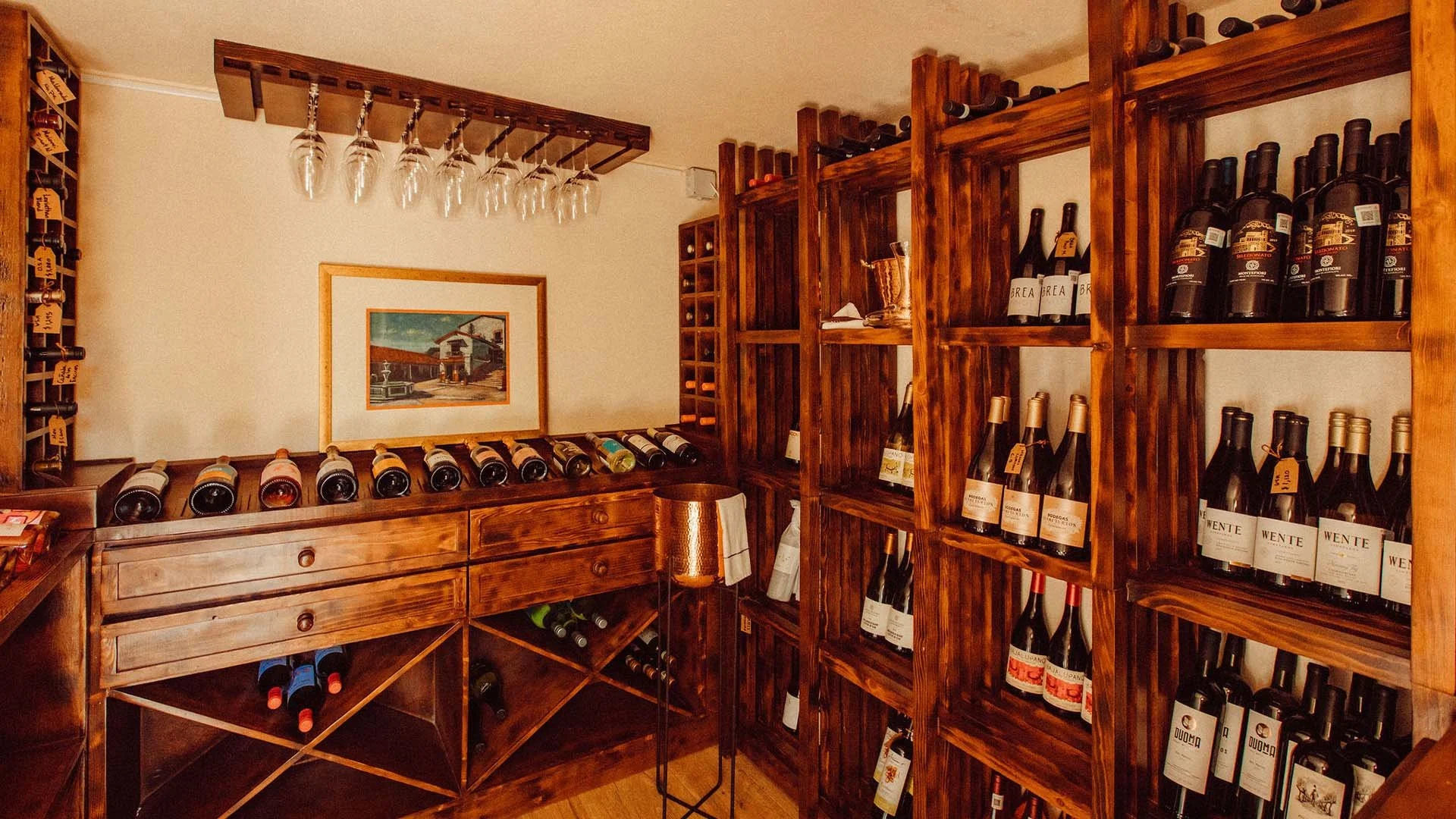 A cozy wine cellar with wooden shelves filled with wine bottles, a rack for hanging glasses, and a wooden cabinet.