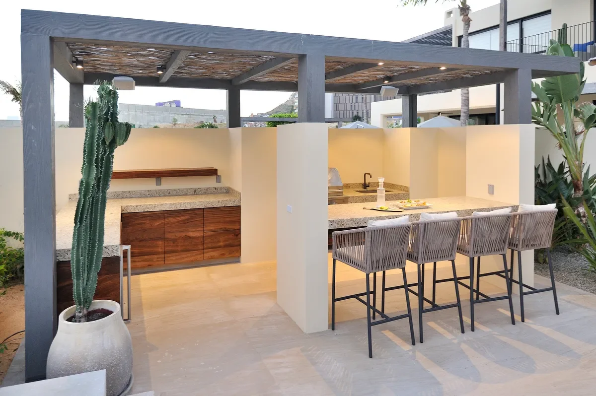Outdoor kitchen with a granite counter, bar stools, and a tall cactus in a pot, set under a wooden pergola in a modern backyard.