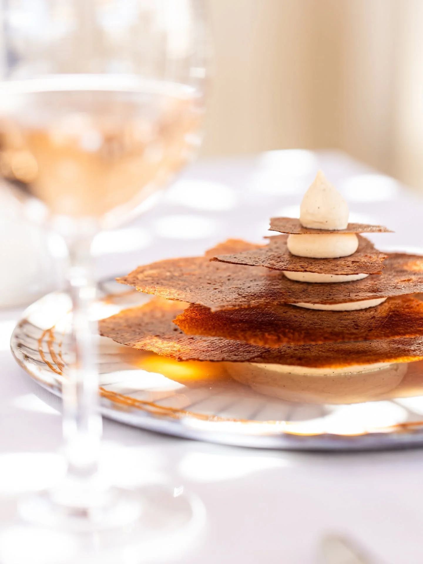 Stacked, crispy crepes with dollops of cream or filling layered between, garnished with a thin chocolate or caramel strip on the top crepe, on a decorative silver tray, with a blurred glass of beverage in the background.