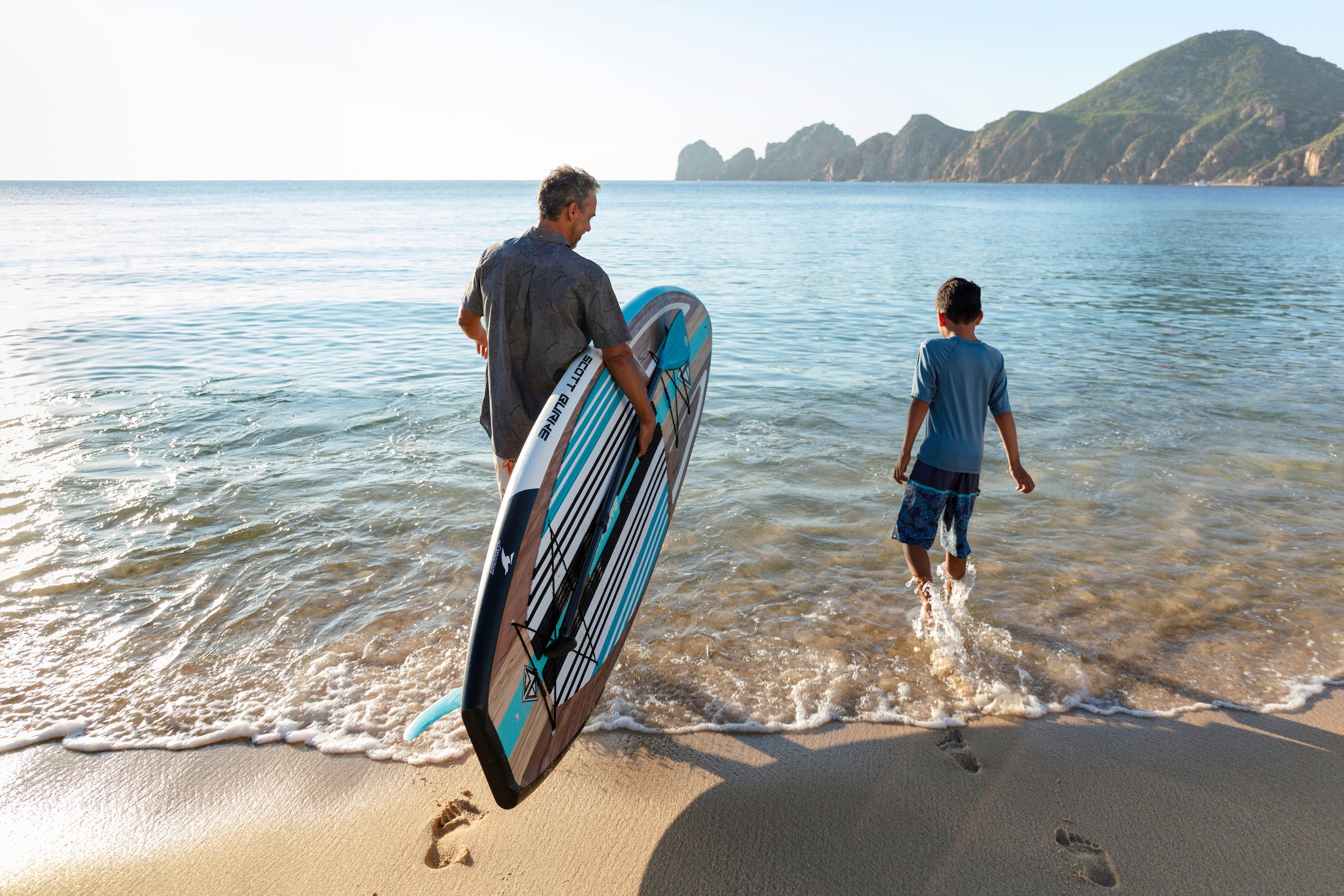 A man and a boy at the beach, with the man holding a paddleboard, walking into the water, and the boy walking ahead, with mountains in the background.