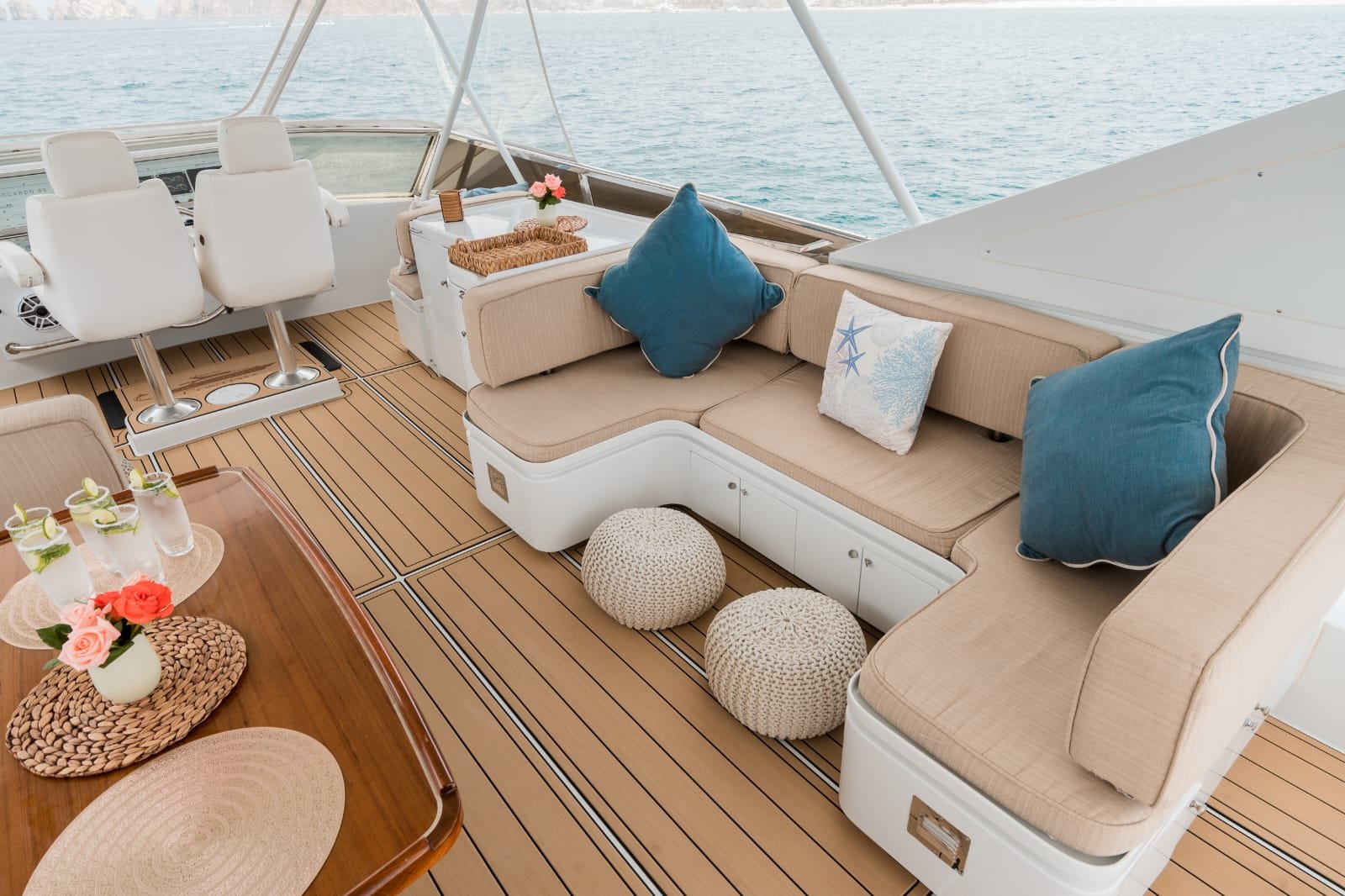Luxury boat deck with beige cushioned seating, blue pillows, woven poufs, and a wooden table with floral decorations, overlooking calm ocean waters.