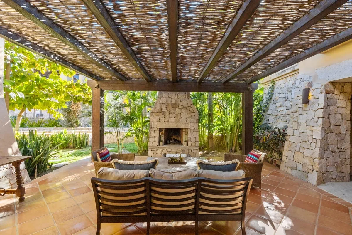 Outdoor patio with a stone fireplace, surrounded by greenery, with a seating area including a sofa and chairs under a wooden pergola.