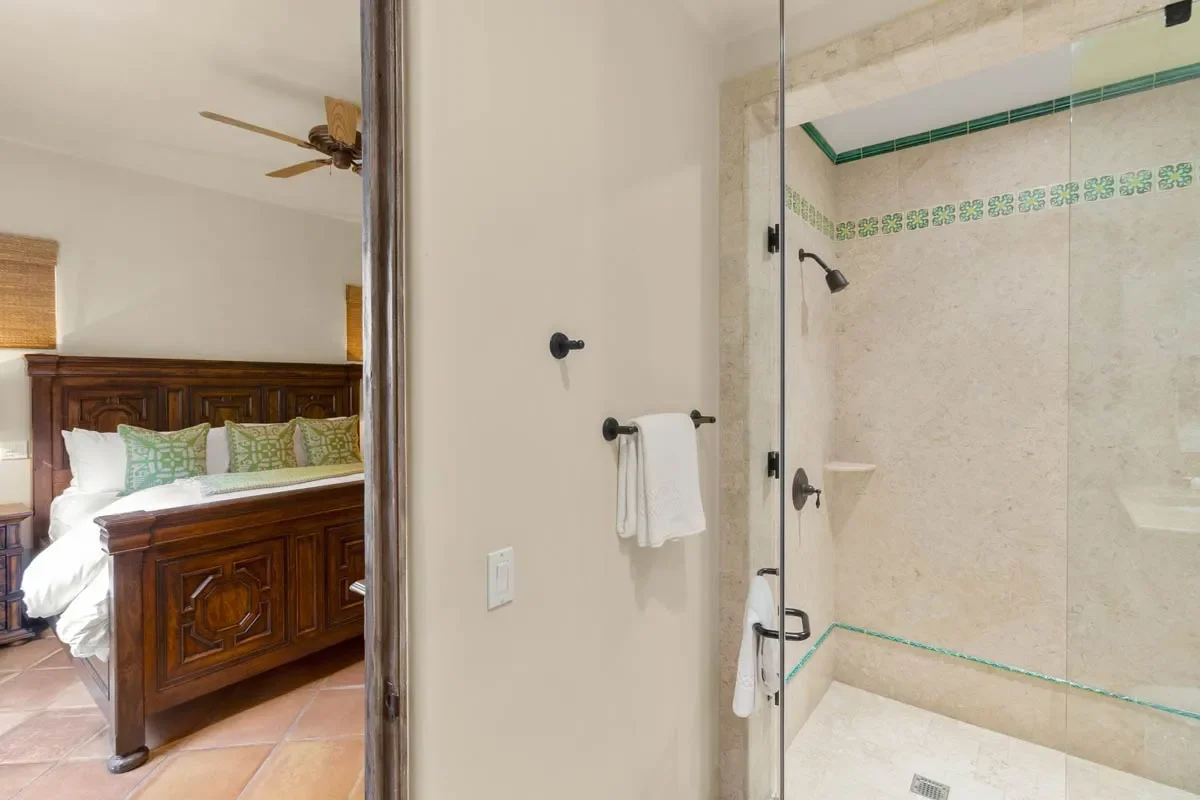 View of a bathroom shower with glass door and towel rack, next to a bedroom with wooden bed and green pillows.