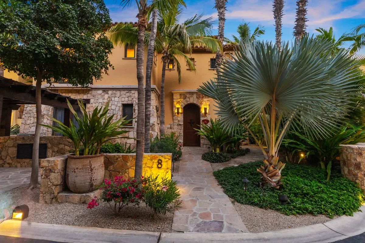 A tropical villa with a stone path leading to a wooden front door, surrounded by lush greenery, palm trees, and decorative plants, with warm lighting illuminating the entrance at dusk.