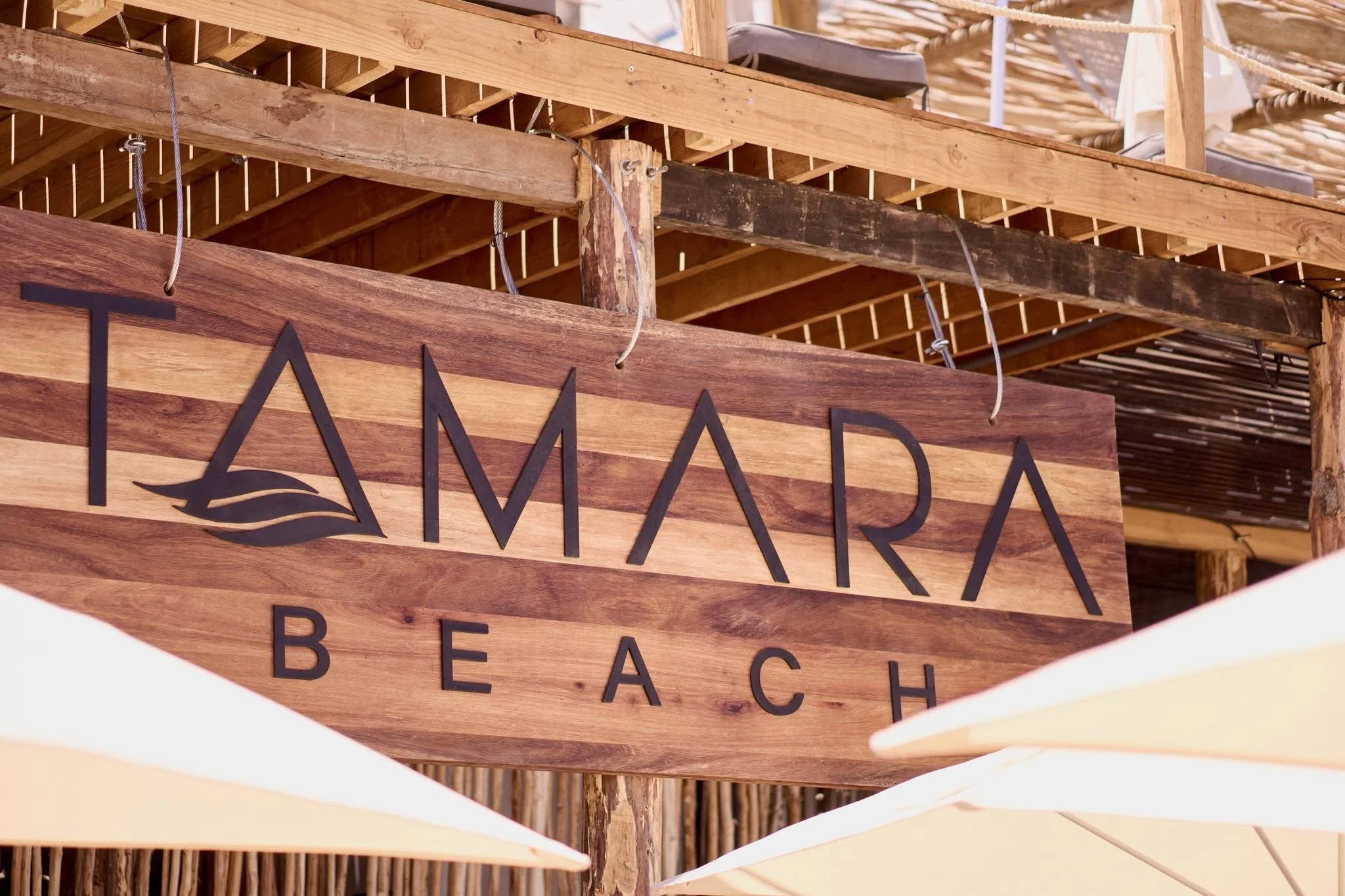 Close-up of a wooden sign that says 'TAMARA BEACH' with a leaf logo, hanging in front of a wooden structure and umbrellas at the beach.