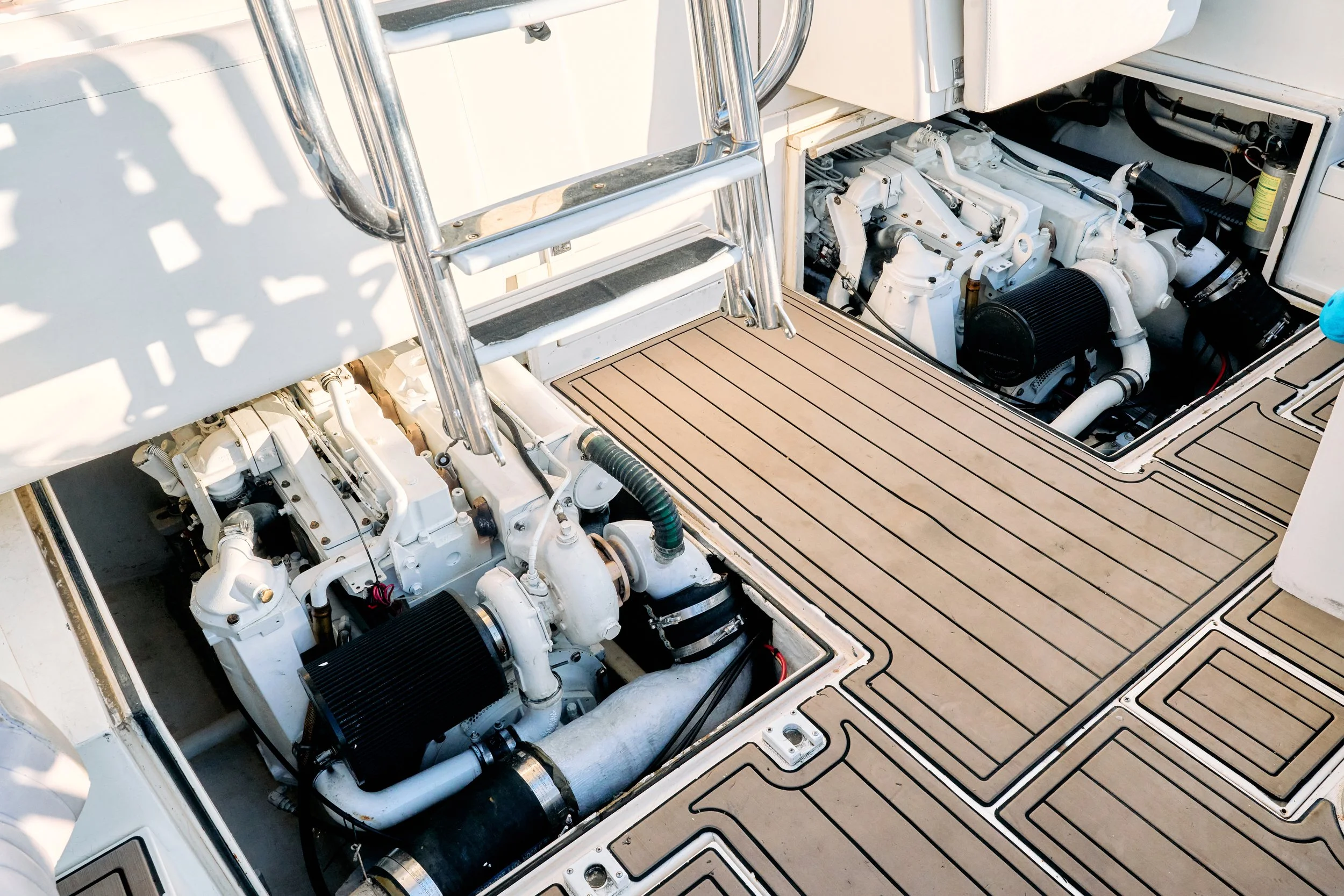 View of a boat's engine compartment with a white engine, black filter, and surrounding pipes, hoses, and cables. There is a metal ladder leading to the upper part of the boat and a wooden platform with a textured surface.