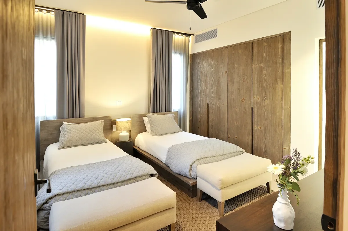 A hotel room with two single beds, wooden accent wall, large windows with gray curtains, a small bedside table with a lamp, a wardrobe, and a vase of flowers on the dresser.
