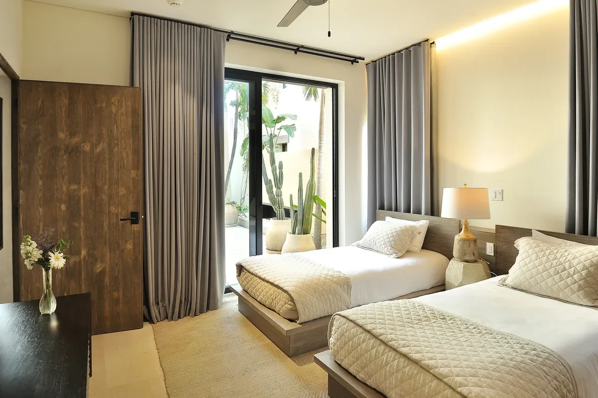 Two beds with white quilts and pillows in a modern bedroom, with a large sliding glass door leading to an outdoor patio with potted plants, and gray curtains on the windows.