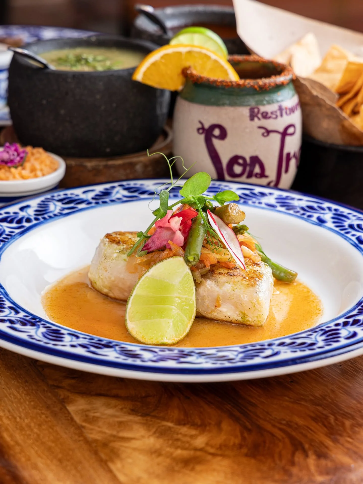 A plated fish dish garnished with lime wedges, fresh herbs, and vegetables in a sauce, on a decorative blue and white plate. In the background are side dishes, a bowl of soup, and a colorful jar with lemon and lime slices.