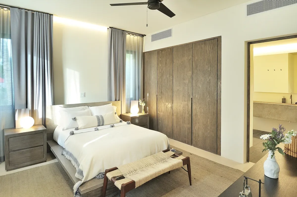 Modern bedroom with a bed, nightstands, drapes, and a closet. Light-colored walls, ceiling fan, and a vanity or dresser with a vase of flowers.