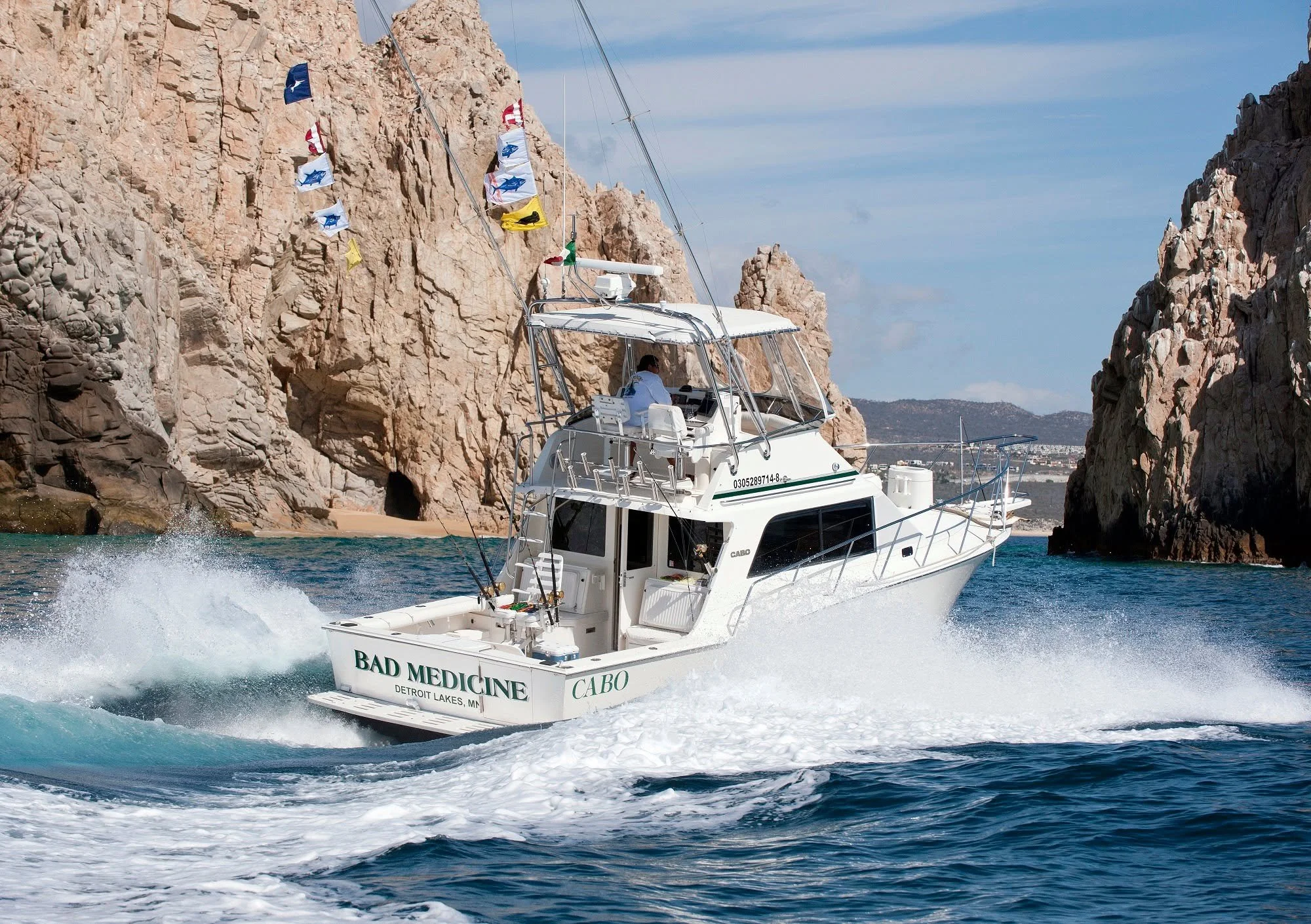 A white yacht named 'Bad Medicine' cruising through blue waters near rocky cliffs under a partly cloudy sky, with flags flying from the mast.