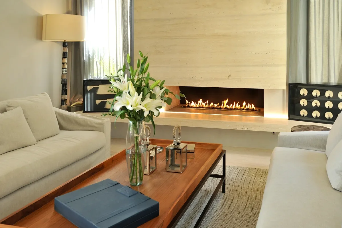 A cozy living room with a fireplace, two beige couches, a wooden coffee table with a flower vase and lanterns, and a tall lamp beside the window.