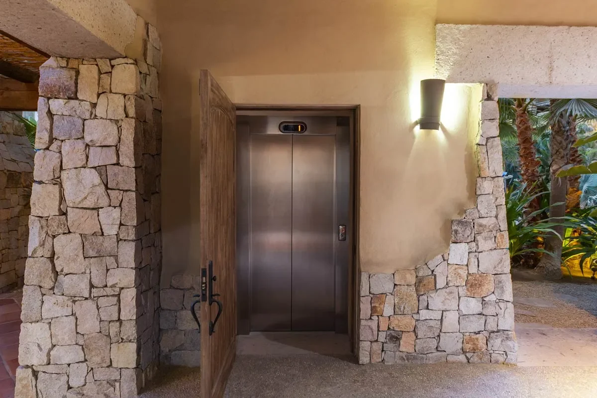 Closed elevator behind partially open wooden door, set in stone wall with outdoor plants and warm wall-mounted light.