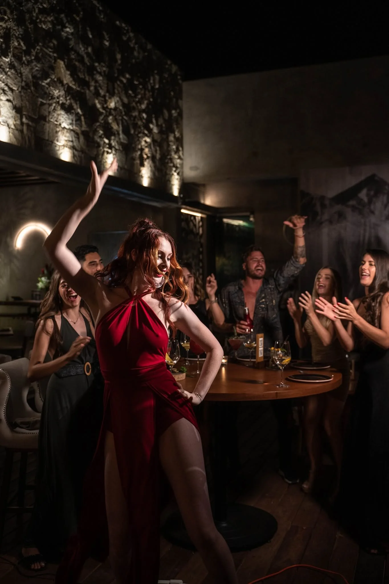 A woman in a red dress dancing at a lively social gathering or party. Several people around her are watching and clapping, enjoying the moment. The setting appears to be indoors with dim lighting and decorative wall features.