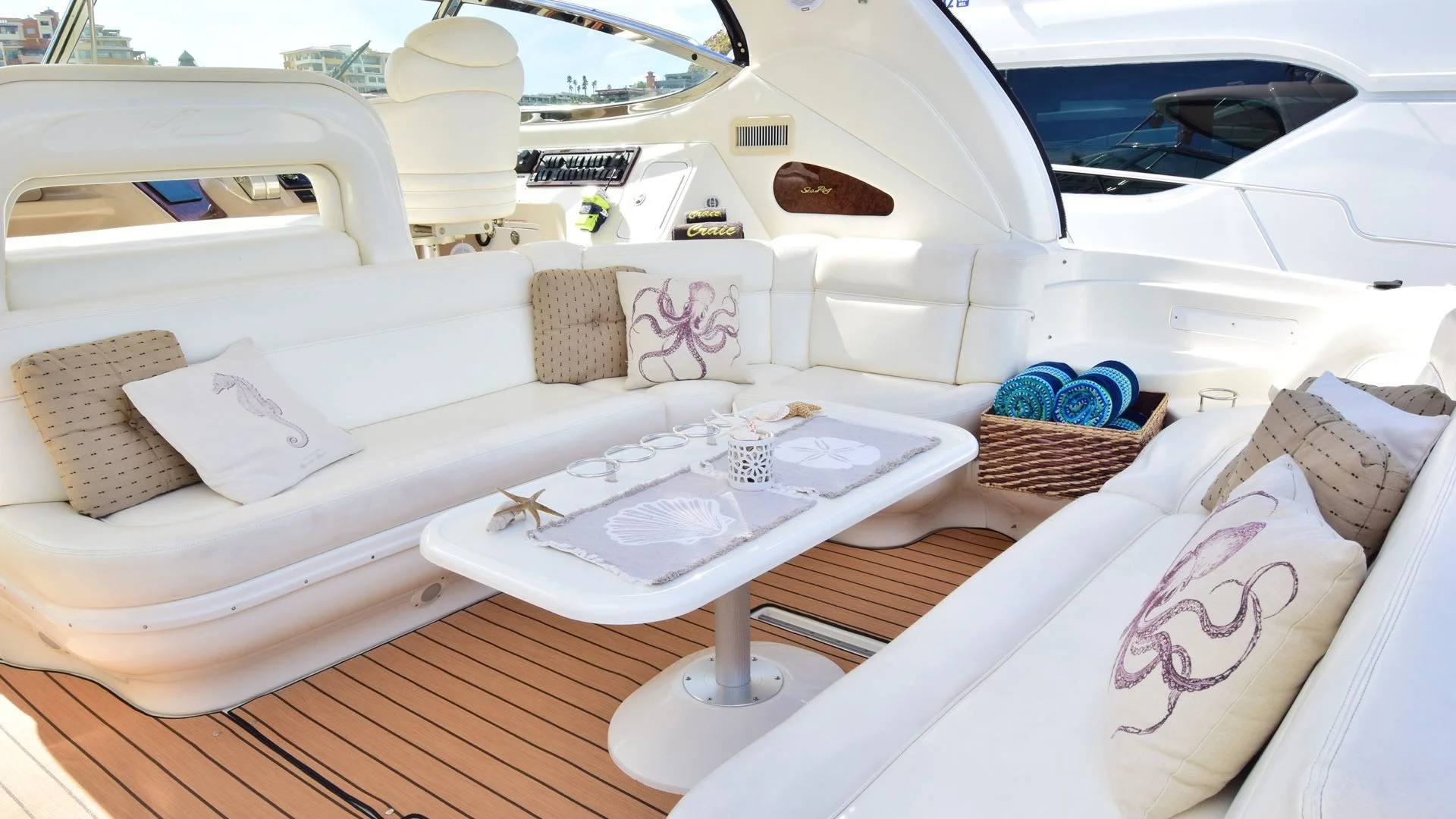Outdoor seating area on a yacht with white cushioned couches, decorative pillows with octopus and seahorse prints, a white table with sea-themed placemats, a basket of rolled beach towels, and a view of a marina with buildings in the background.