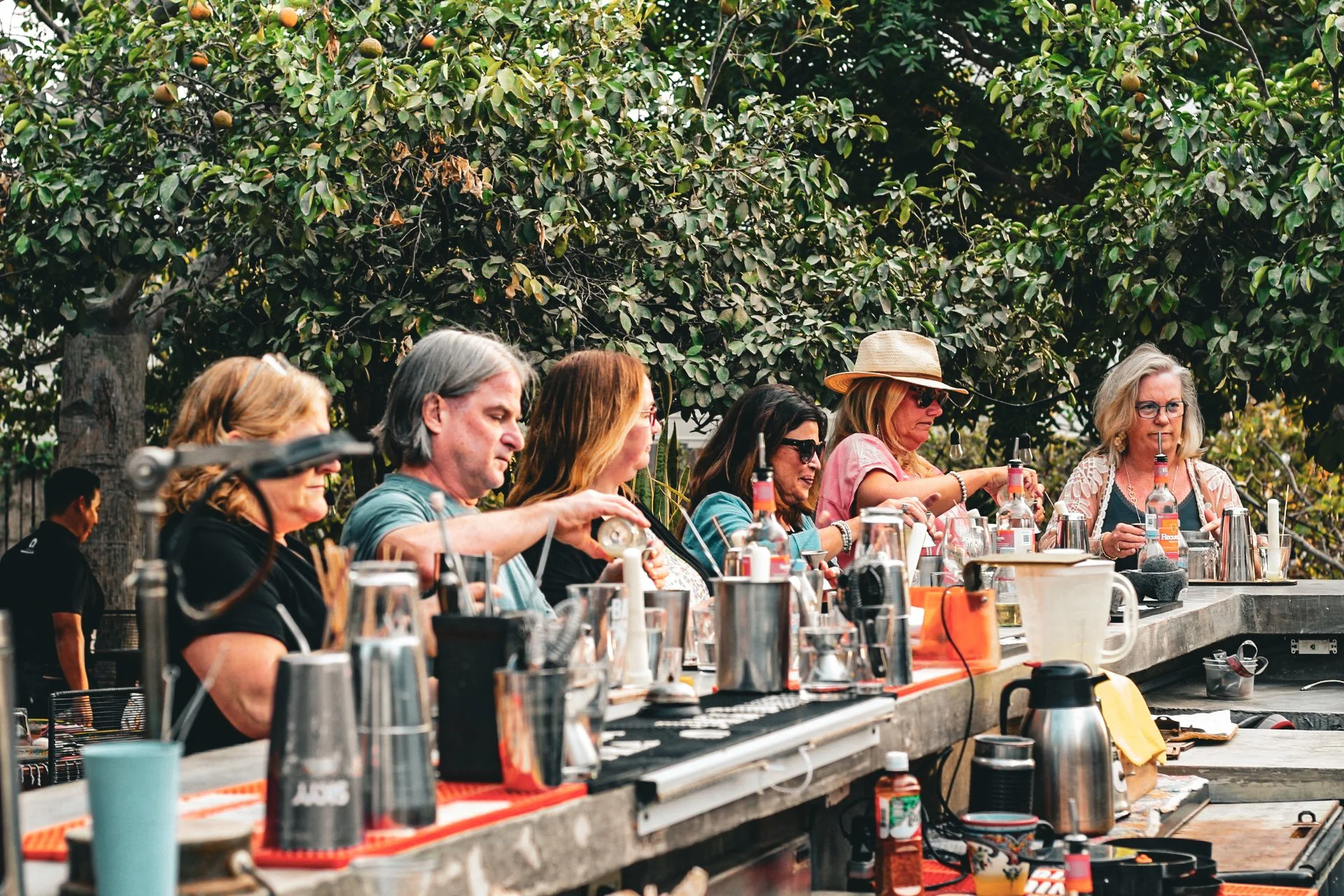 People gather outdoors around a bar counter with various bottles and barware, under trees with green leaves and some fruit, possibly in a park or garden setting.