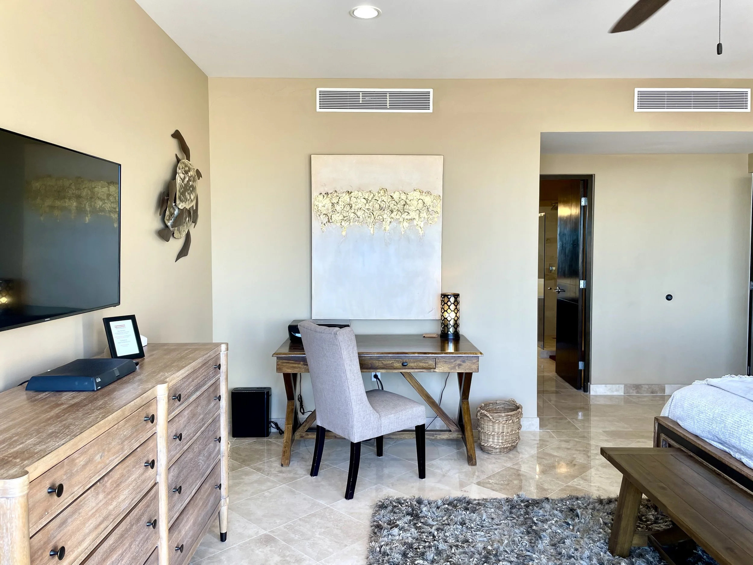 A living room with beige walls and a white ceiling. There's a flat-screen TV on the left wall, a wooden dresser below it, and various decorative items including a small framed picture and a black speaker. In the center, a wooden desk with a gray upholstered chair is against the wall, with a tall lamp on it. Above the desk, a large abstract painting with neutral colors is hung. To the right, part of a yellowish-beige armchair and a wooden bench are visible, with a textured gray and black area rug on the tiled floor. The ceiling has two vents and a ceiling fan with a pull chain.