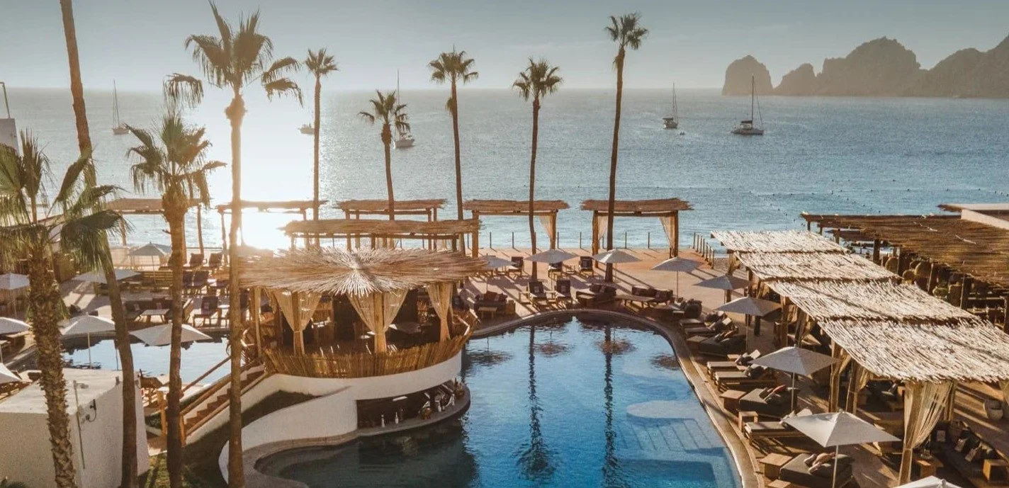 Beachside resort with a swimming pool, palm trees, lounge chairs with umbrellas, shaded cabanas, and boats on the ocean in the background at sunset.